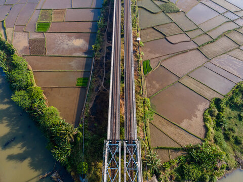 Aerial view of a railway bridge crossing over lush rice fields and a river in Banyumas Regency, Central Java, Indonesia.