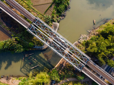 Aerial view of a steel railway bridge crossing a river, surrounded by lush tropical vegetation and agricultural fields in Banyumas Regency, Central Java, Indonesia.
