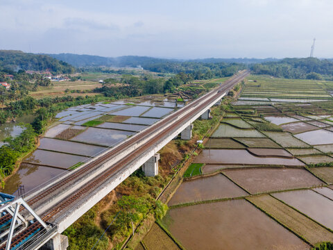 Aerial view of a modern railway bridge crossing over flooded rice paddies in a rural landscape with lush hills in the background in Banyumas Regency, Central Java, Indonesia.