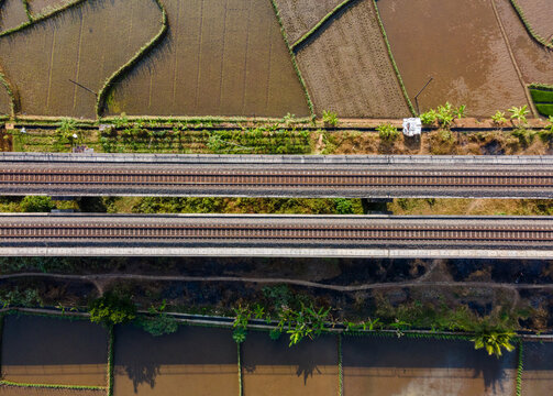 Aerial view of a double-track railway bridge crossing over lush green and brown rice fields in the rural landscape of Banyumas Regency, Central Java, Indonesia.
