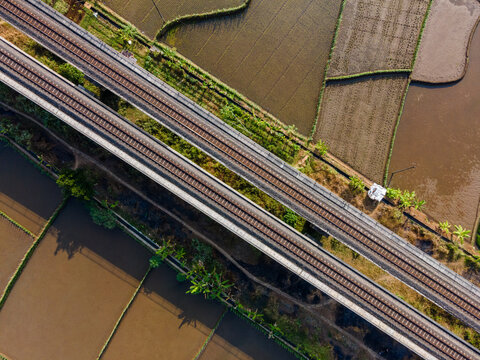 Aerial view of a double-track railway bridge spanning across lush green and brown rice paddies in the rural landscape of Banyumas Regency, Central Java, Indonesia.