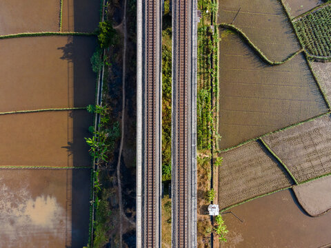 Aerial view of a double railway track cutting through lush green and brown rice paddies in the rural landscape of Banyumas Regency, Central Java, Indonesia.
