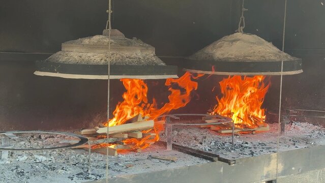 Open flame cooking station with two large metal lids over a charcoal hearth, bright orange fire and ash create a rustic kitchen mood for traditional food prep