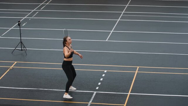Woman practicing badminton on court, solo training with powerful smashes and focused serves, black athletic wear, tripod camera in background, deliberate footwork and rhythm drills for strength