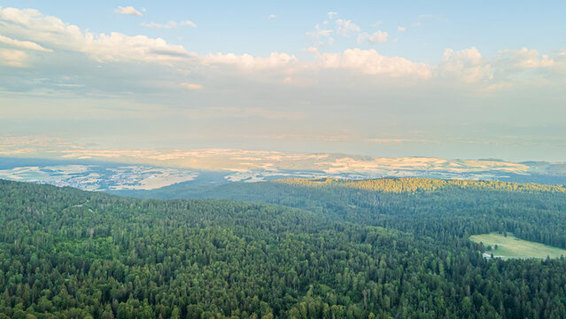 Aerial view of the Jura mountains landscape with dense green forests, rolling hills, and distant plains under a soft sky in Jura, Switzerland.