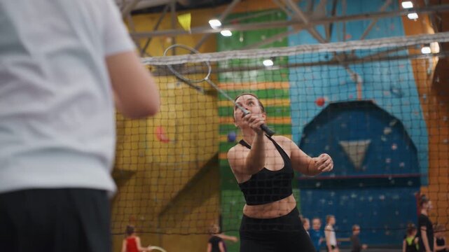 Woman athlete returning shuttlecock during intense badminton rally on indoor court with partner opposite and climbing wall backdrop, expressive reactions, paced volleys and attacking smashes,