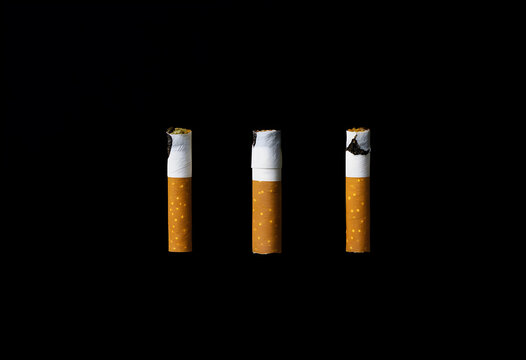 Three lit cigarettes with burning tips and ash against a dark black background, addiction and health risk