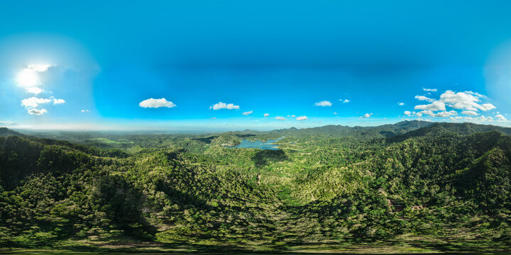 Aerial view of Sermo Reservoir Dam surrounded by lush green hills and dense forest under a clear blue sky in Kecamatan Kokap, Daerah Istimewa Yogya, Indonesia.