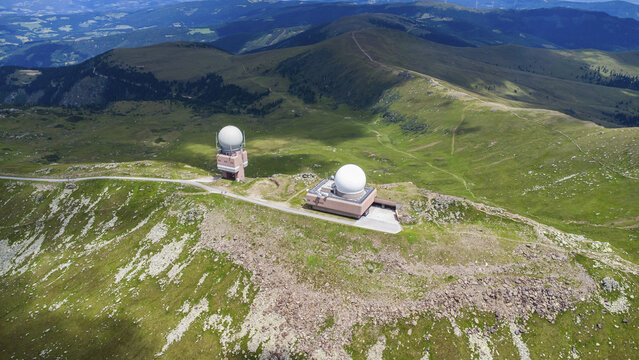 Aerial view of the Koralpe radar station with two white radomes on a mountain ridge surrounded by green alpine slopes and rocky terrain in Wolfsberg, Carinthia, Austria.