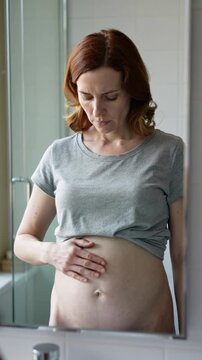 Pregnant woman in grey t-shirt touching belly in front of bathroom mirror, eye level shot