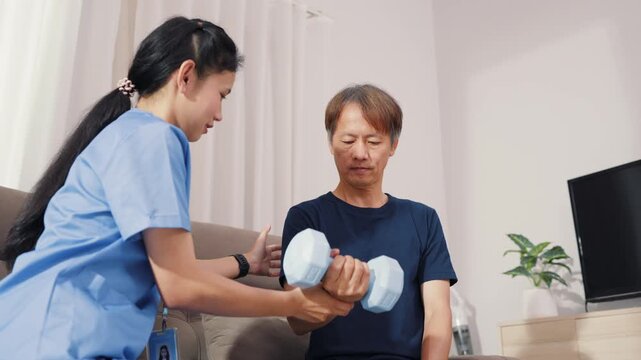 Supportive Asian therapist assisting male patient with dumbbell lifting exercise for physical therapy. Dedicated healthcare professional guiding strength recovery training on sofa.