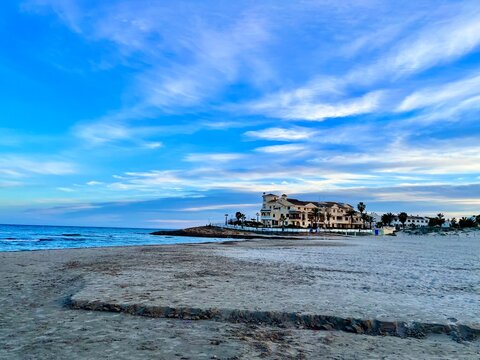Amplia playa de arena fina frente a complejo residencial costero bajo un din&aacute;mico cielo con nubes