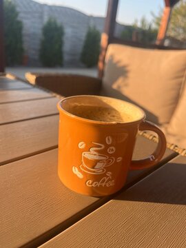 Taza de caf&eacute; naranja con espuma sobre mesa de madera en una terraza durante el amanecer