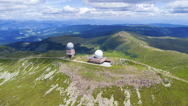 Aerial view of the Koralpe radar station with its distinctive white domes set amidst the lush green alpine meadows and rocky slopes of the mountain range in Wolfsberg, Carinthia, Austria.