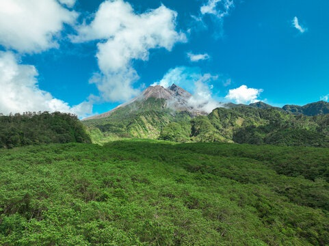 Aerial view of Merapi Mountain rising above lush green tropical forests under a bright blue sky with scattered clouds in Kecamatan Cangkringan, Daerah Istimewa Yogya, Indonesia.