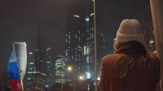Night skyline view from promenade woman in knit hat watches illuminated highrises and flag display, soft streetlights, reflective glass, chilly air, traveler taking in urban panorama