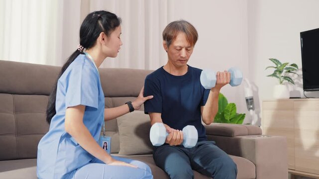 Supportive Asian therapist assisting male patient with dumbbell lifting exercise for physical therapy. Dedicated healthcare professional guiding strength recovery training on sofa.