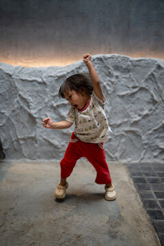 Energetic Asian toddler girl dancing and jumping with raised hands against textured concrete wall.