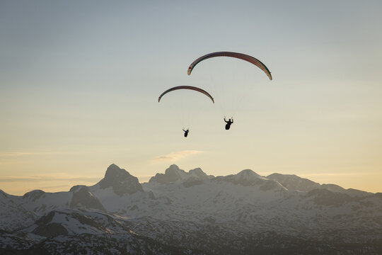 Aerial view of two paragliders soaring above the snow-capped peaks of the Dachstein massif during golden hour in Grobming, Styria, Austria.