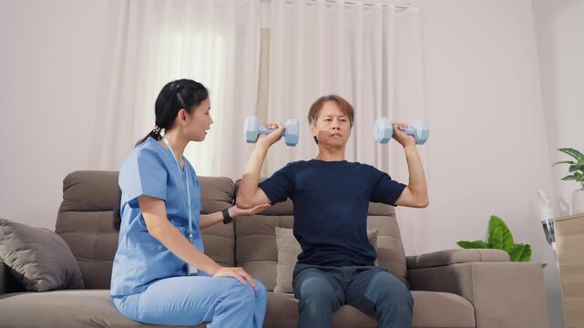 Supportive Asian therapist assisting male patient with dumbbell lifting exercise for physical therapy. Dedicated healthcare professional guiding strength recovery training on sofa.