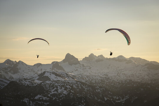 Aerial view of paragliders soaring above the snow-capped peaks of the Dachstein mountains during golden hour in Grobming, Styria, Austria.