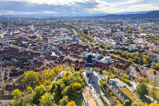 Aerial view of the Schlossberg hill with the iconic Clock Tower, historic red-roofed buildings, the Mur river, and autumn trees in Graz, Steiermark, Austria.