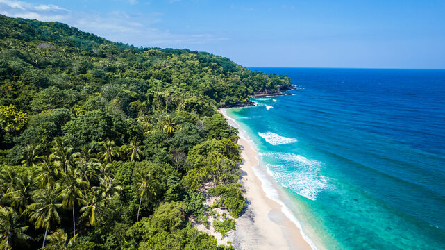 Aerial view of Waebela beach featuring lush tropical forest, turquoise water, and gentle waves hitting the sandy shore in Inerie, East Nusa Tenggara, Indonesia.