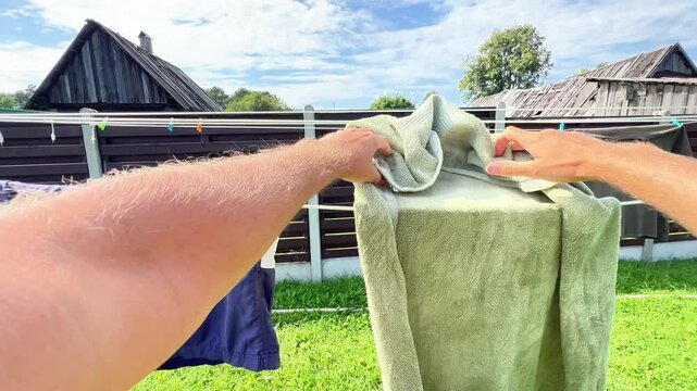 First-person view of a person securing wet laundry and towel with colorful plastic clothespins on an outdoor clothesline in a sunny garden during summertime.