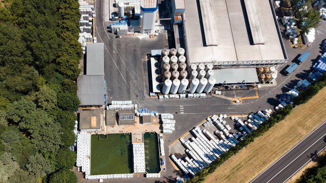 Aerial view of the Mapei industrial facility with large storage silos, warehouse buildings, and outdoor inventory areas surrounded by trees and a nearby road in Mediglia, Lombardia, Italy.