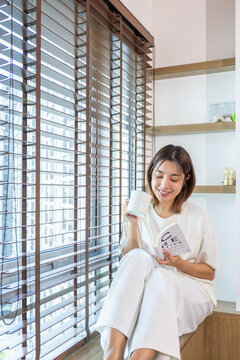 Asian woman holding a mug and reading a book beside the window