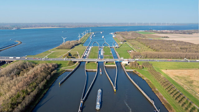 Aerial view of the Volkerak locks with cargo ships navigating the waterway, surrounded by green embankments, wind turbines, and a highway bridge in Willemstad, Noord-Brabant, Netherlands.
