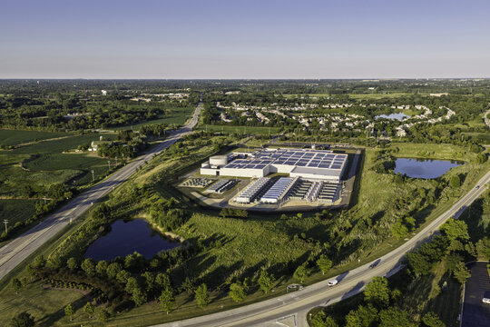 Aerial view of a data center facility with solar panels on the roof, surrounded by green fields, ponds, and roads in Illinois, United States.
