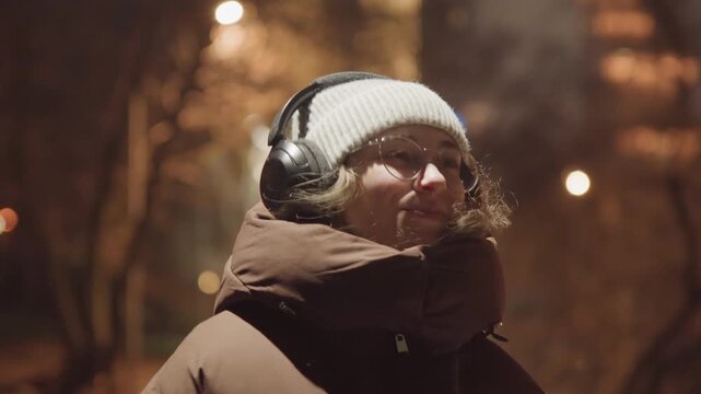 Young woman listening to music on quiet park path, closeup portraits show blissful expressions and playful pouts under warm streetlight bokeh, cozy beanie and thick coat emphasize winter warmth
