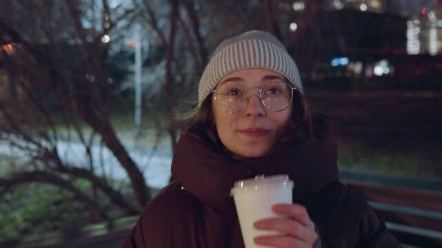 Young white woman on bench holding coffee cup, wearing knit beanie and glasses, puffer jacket, soft city lights and bokeh in background, visible breath in cold air, gentle smile, candid evening