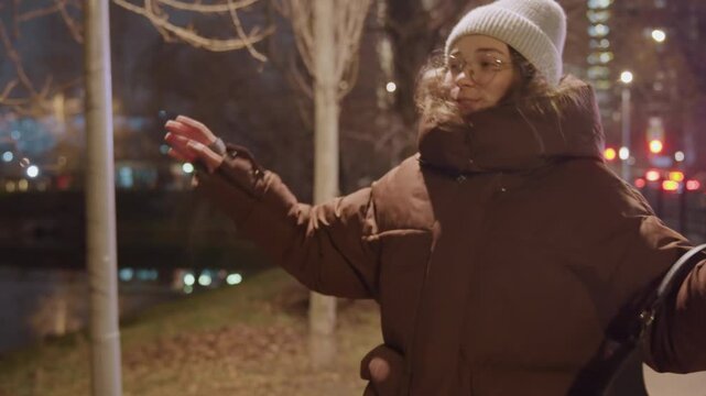Young woman moves lively in city night. Energetic young woman dances under streetlights at night. Bright city illumination highlights woman dancing cheerfully in winter clothing during nighttime