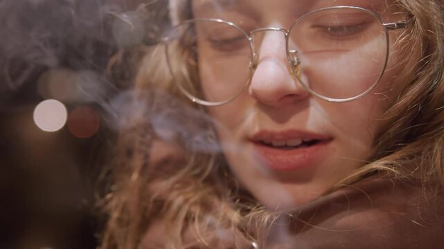 Subtle exhale captures melancholic evening mood. Intimate face illuminated by city glow and soft smoke drifting. Serene latenight scene showing person with gentle smoke and reflective mood