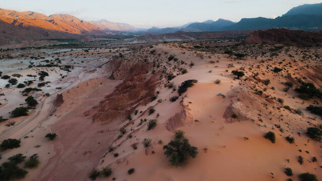 Aerial view of Valles Calchaquies featuring rugged red rock formations, desert dunes, sparse vegetation, and distant mountains under soft golden hour light in San Carlos, Salta, Argentina.
