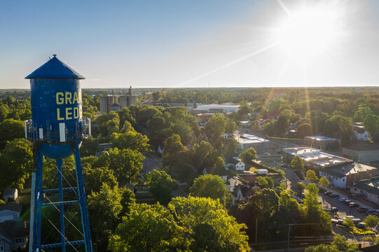 Aerial view of the blue Grand Ledge water tower rising above lush green trees and residential streets in the golden hour light in Grand Ledge, Michigan, United States.