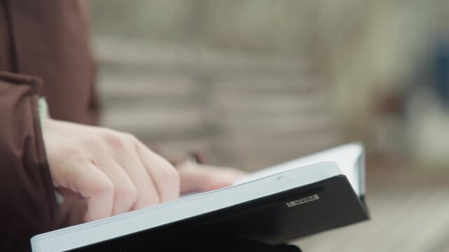 Closeup hands flipping pages of planner on bench, brown coat, pen poised, organized layout, purposeful posture urban planner scheduling goals, neat handwriting, overcast park background, steady