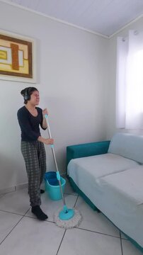Happy woman wearing headphones dancing and singing energetically while mopping the floor