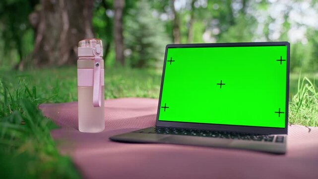 Laptop with green screen and tracking markers sitting on yoga mat next to water bottle in park. Mockup template is ideal for promoting online fitness classes, wellness apps or remote training
