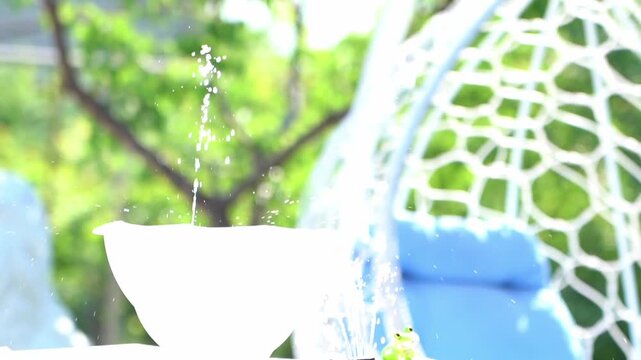 White bowl catches falling water droplets. Egg-shaped chair features a blue cushion. Lush green trees and a house appear behind. Garden scene offers calm, peaceful vibes