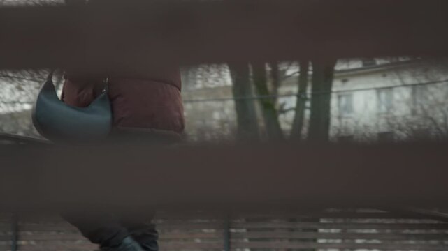 Closeup boots walking across wooden deck, camera peeking through bench slats, partial silhouette and coat hem visible, rhythmic footsteps over cold wood, voyeuristic observational mood