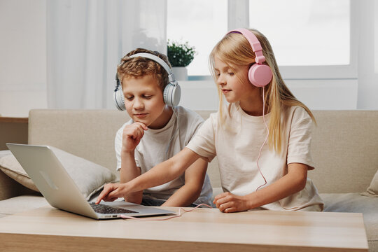 Two children with headphones using laptop on table in cozy living room. Concept of technology, education, and entertainment at home for kids.