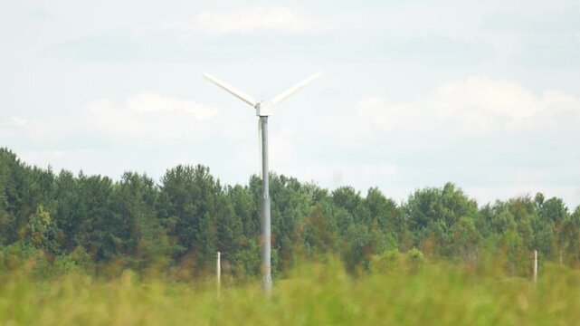 Wind turbine spins steadily in open grassland. Dense forest and partly cloudy sky form the backdrop. Green landscape highlights renewable energy potential