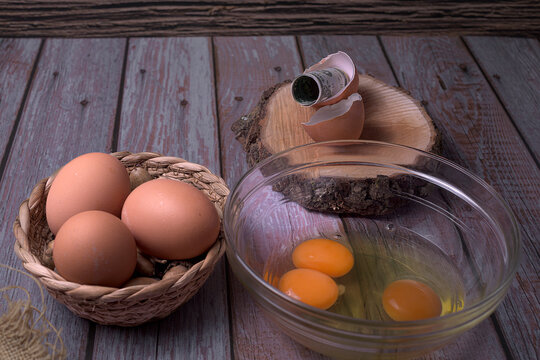 Whole brown eggs in a basket, raw yolks in a glass bowl, and rolled dollar bill inside an eggshell on wooden background