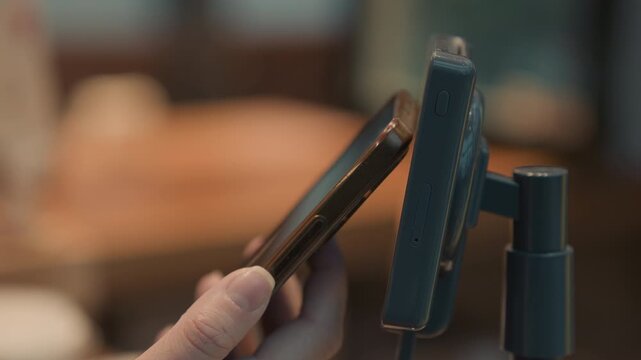 Smartphone tap on card reader terminal, hand holding phone performing contactless payment at cozy cafe counter with blurred bokeh background, mounted reader on adjustable stand, warm lighting