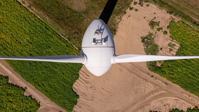 Aerial view of a white wind turbine nacelle and blades positioned above a rural landscape with green fields and sandy soil in Poland.