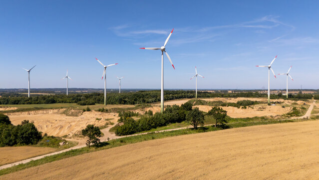 Aerial view of wind turbines standing in a rural landscape with fields, trees, and a sandy quarry under a clear blue sky in Poland.