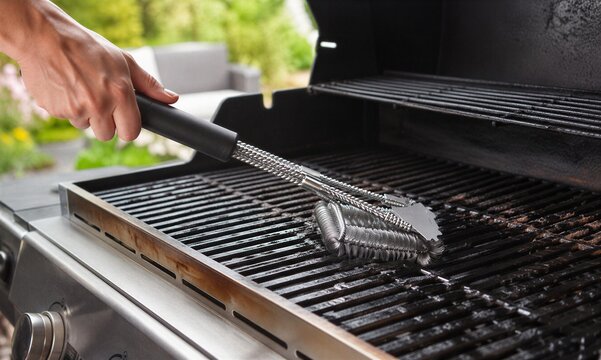 Hand cleaning dirty gas grill grate with bristle-free coil brush on outdoor barbecue, concept of grill maintenance, food safety, seasonal backyard cooking, and household cleaning.
